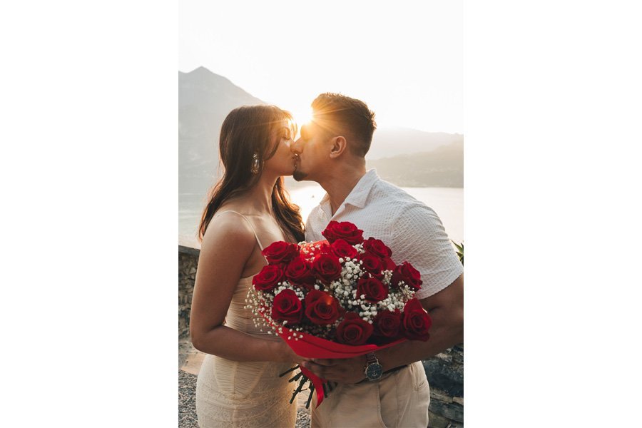 Two New Fiancés Kiss While Hold Red Roses At Sunset in lake como